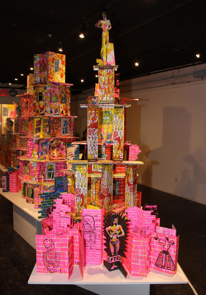 Photo of large paper sculptures on display, the sculptures are arranged in towers, and have various illustrations, textures and colors, including brick patterns, male figures and the cut out windows. 