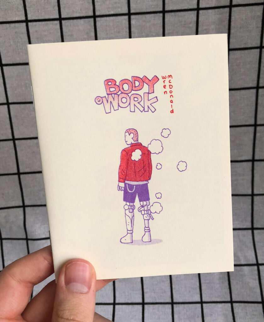 Photo of a small zine titled "Body Work" printed in red and purple, showing a male figure with robotic legs standing and facing away from the viewer, puffs of smoke surround him. 