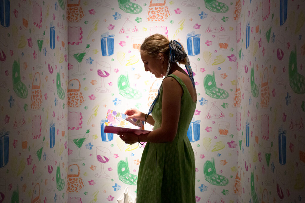 A woman in a green dress with her hair tied up stands facing left in a small space, three walls are covered in a repeating pattern of colorful illustrations of various objects such as purses, wine glasses and cell phones. 