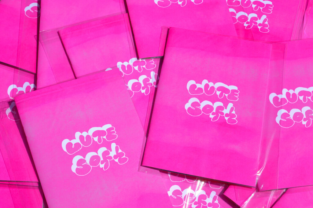 A photograph of many copies of a zine titled "Cute Soft" in bubble letters against a pink background. 