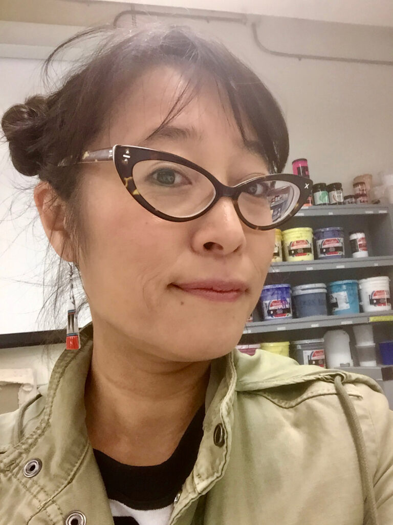 Image of Asuka, who wears her black hair in a bun and cat-eye glasses.
