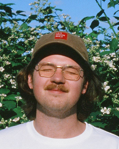 Photo of Bjorn Miner, who wears a baseball cap and glasses, and stands against plants and flowers. He has brown hair down to the base of his neck, a small mustache, and his eyes are closed.