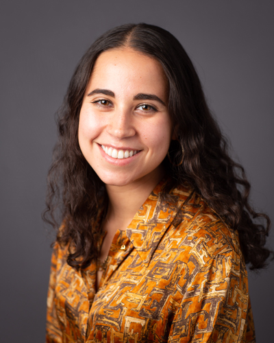 Photograph of Erin Palumbo, who has long dark curly hair, and wears an orange patterned shirt.
