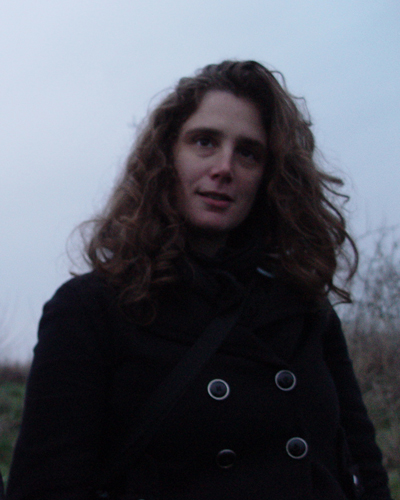 Photograph of Fawn Krieger, who smile slightly and has deep set eyes and long curly hair. She wears a black coat and stands against a cloudy grey sky.