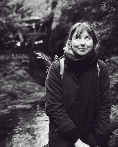 Black and white photo of Natalie who looks to her right, wearing a scarf and backpack, standing in front of trees and water.