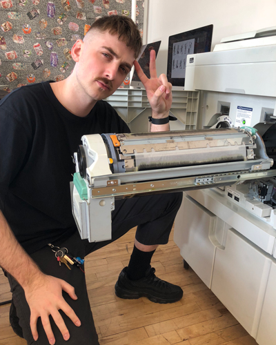 Photo of Wren McDonald kneeling next to a riso drum that's pulled out in the machine, wearing black and holding up a peace symbol.