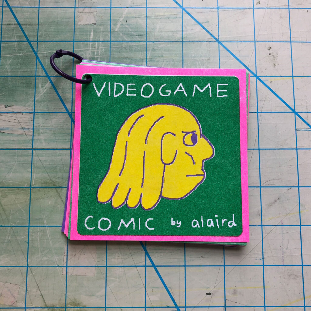 Photo of a small book titled "Videogame" the cover shows a head facing right in yellow, against a green background with a pink border. It is bound by a single plastic ring in the left corner. 