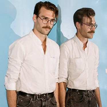 Photograph of Aidan Fitzgerald, who wears glasses, and has a mustache and a swoop of hair down the left side of his face, standing next to a mirror.