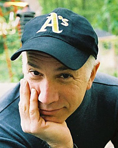 A photo of Stewart Wilson, who leans his chin in his hand. He wears a dark baseball cap and a dark shirt.