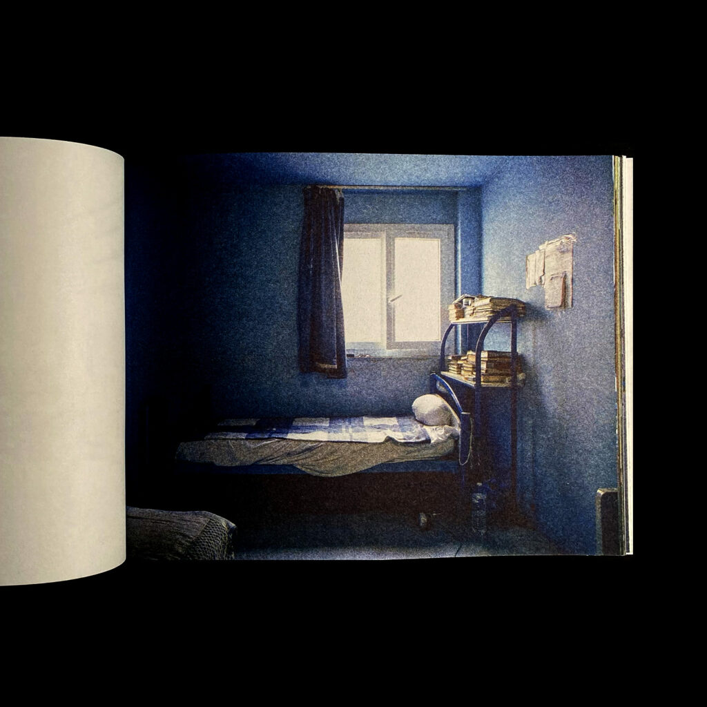 Open spread of a riso photograph of a bedroom, facing a window and twin size bed with a shelf of books behind it, mostly blue and black tones.
