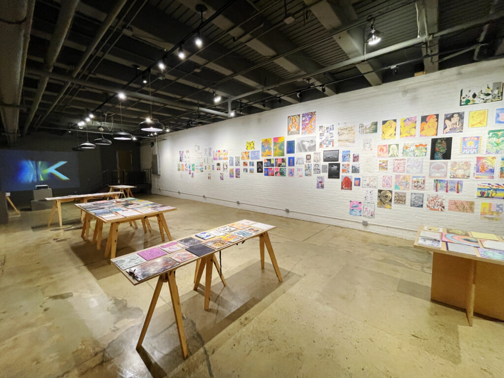 Photo of "Printing the Future" exhibition, showing zines laid out across multiple wooden tables and prints arranged on the walls salon style. A projector plays an animation against the far wall.