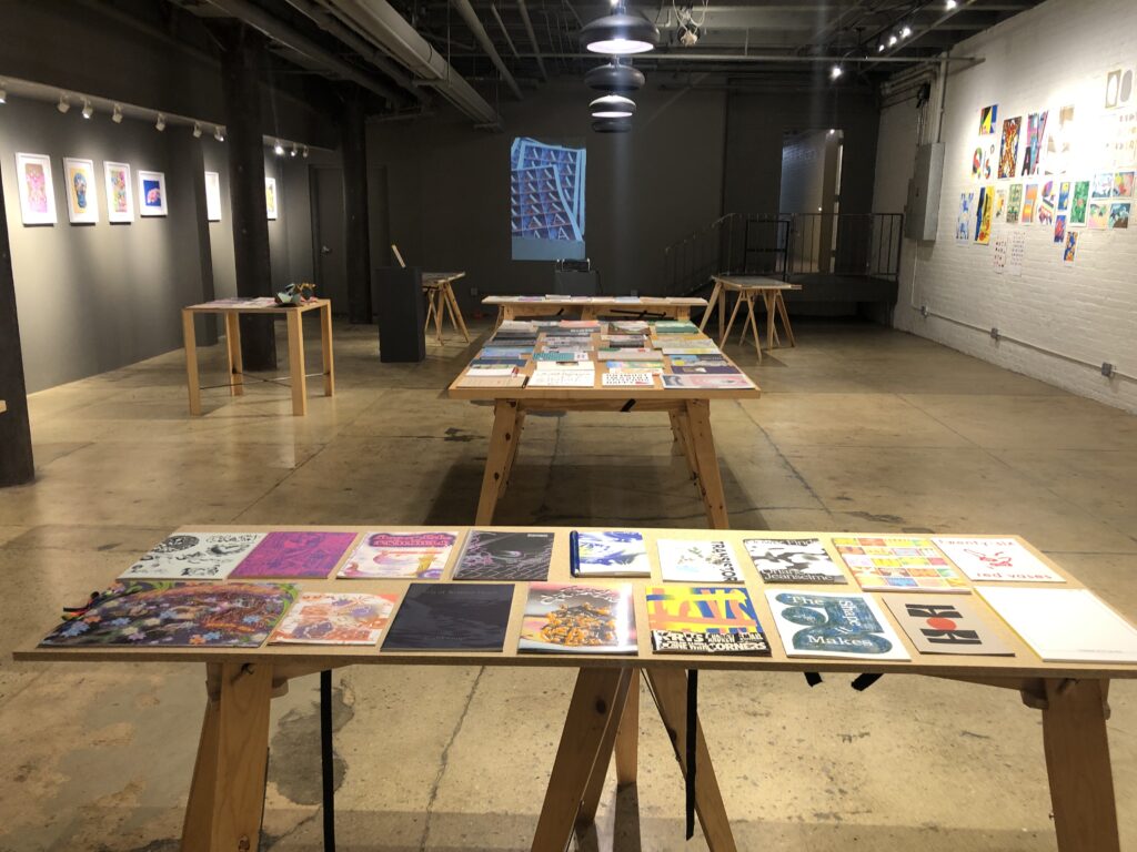 Photograph of Printing the Future Exhibition, riso prints line the walls and zines and books are arranged on wooden tables. A riso animation is projected on the back wall of the room.