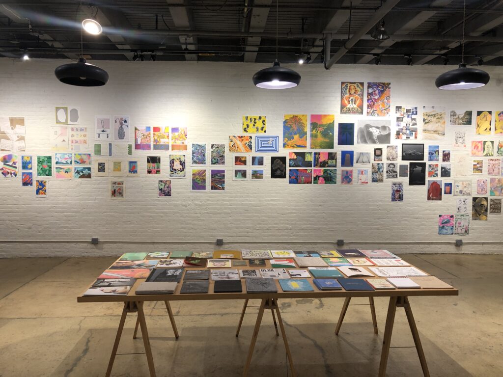 Photograph of Printing the Future Exhibition, riso prints line the walls and zines and books are arranged on wooden tables.