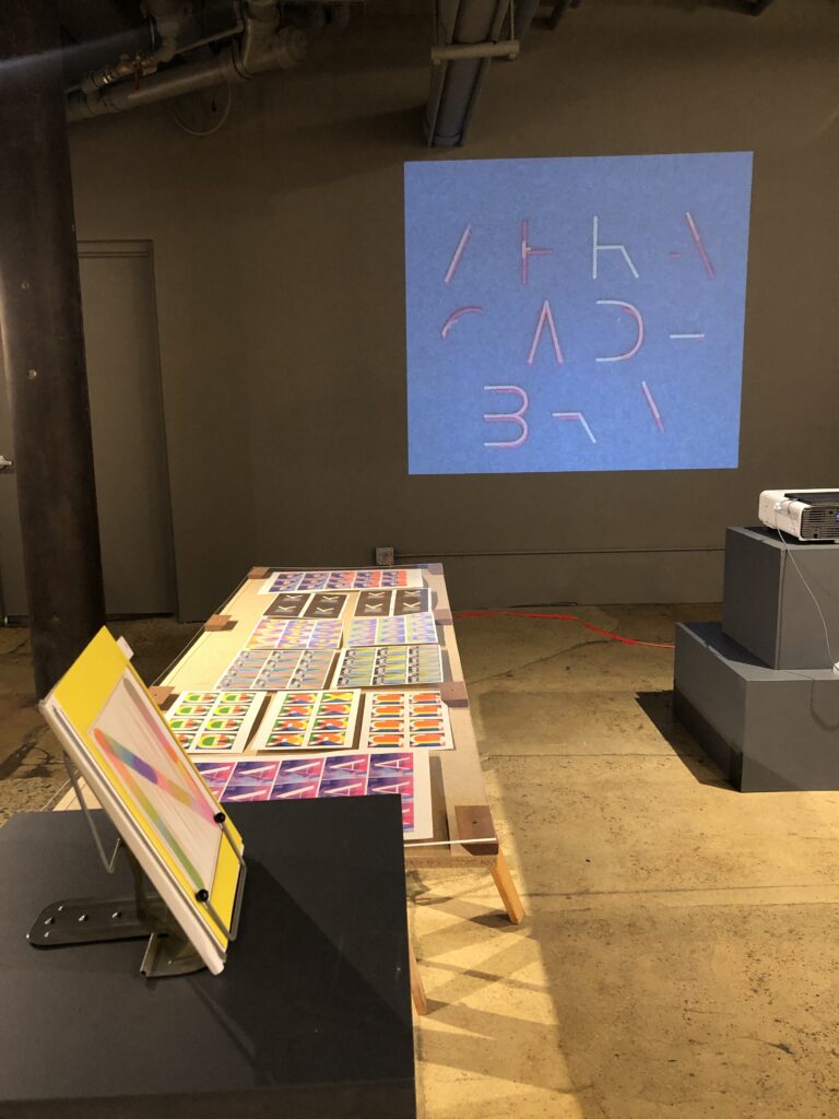 Photograph of Printing the Future Exhibition, zines and books are arranged on wooden tables. A riso animation is projected on the back wall of the room.