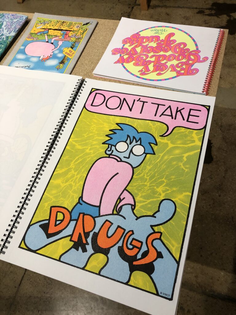 Photograph of riso books on a table, the closest spread shows an illustration of a blue figure looking at us over their shoulder, reaching a hand toward us, holding big orange letters that spell "DRUGS" against a green water texture background. A pink speech bubble reads "DON'T TAKE" across the top.