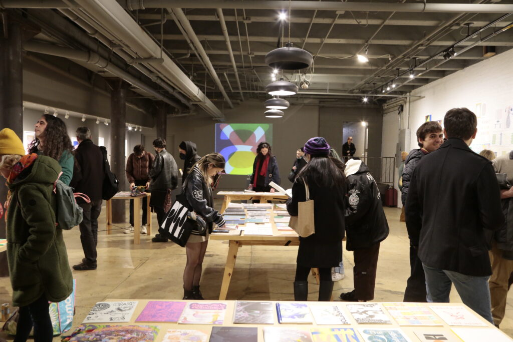 Photo of opening night at "Printing the Future" exhibition, showing zines laid out across multiple wooden tables and prints arranged on the walls salon style. Many people crowd around looking at the artwork.