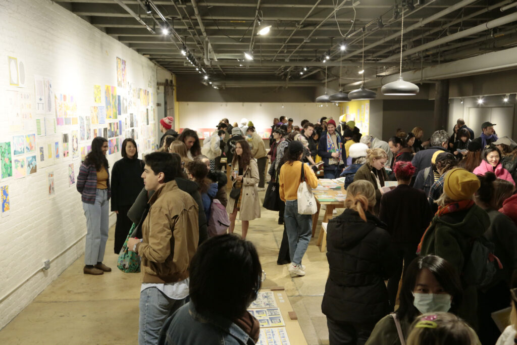 Photo of opening night at "Printing the Future" exhibition, showing zines laid out across multiple wooden tables and prints arranged on the walls salon style. Many people crowd around looking at the artwork.