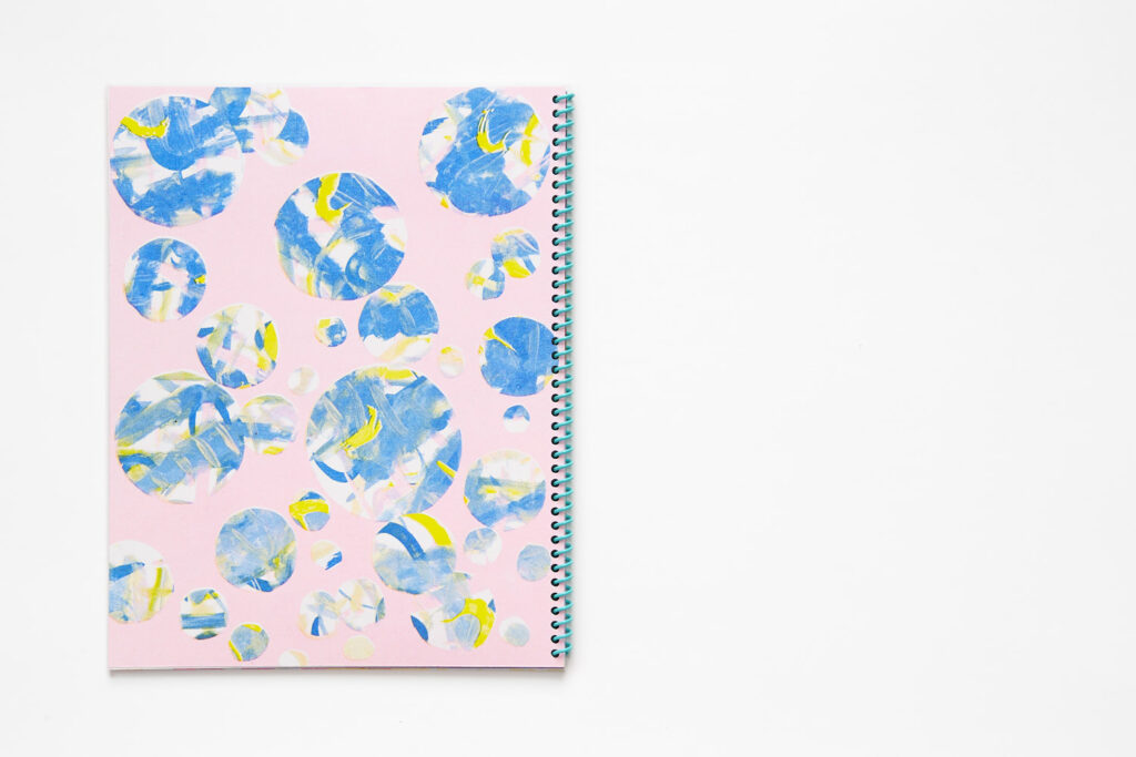 Spiral bound book with an aqua coil, the back cover showing circles of varying sizes with blue and yellow textures and patterns against a light pink background.