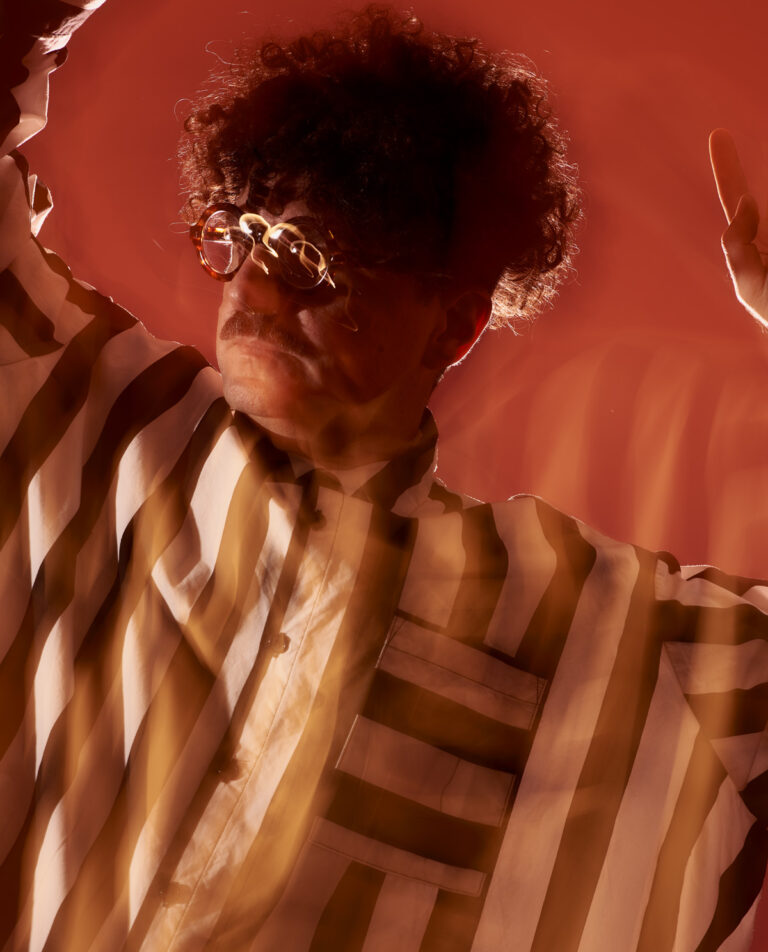 Warm, slighted distorted photograph of risoLAB AIR Marco Scozzaro, who wears a black and white striped shirt, thick framed circular glasses, and has curly hair and a mustache.