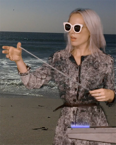 Photograph of Kacie Lees standing on the beach, with dyed gray hair, thick sunglasses and a textured jacket.
