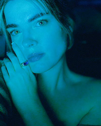 Photograph of Kate Hush, holding a hand next to her face, covered in blue light.