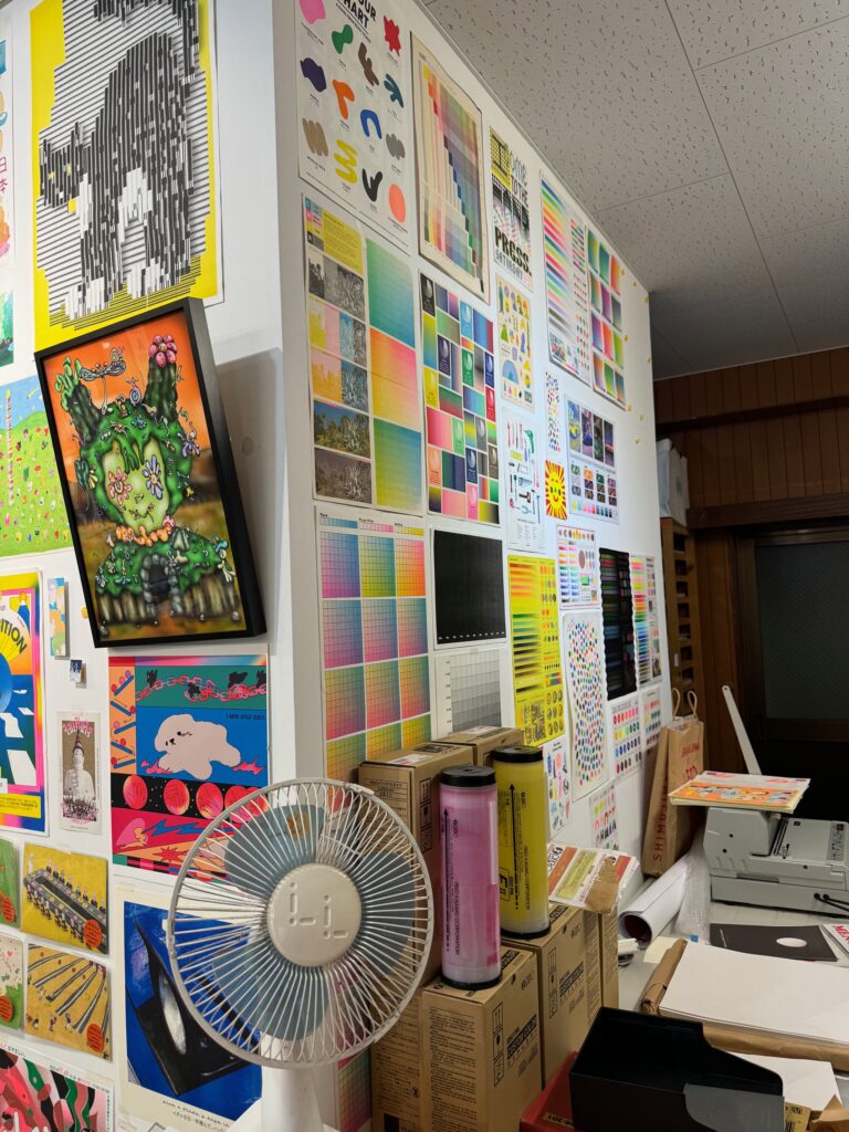 INterior studio view showing a wall of color charts, a fan in the forground, prints covering every surface