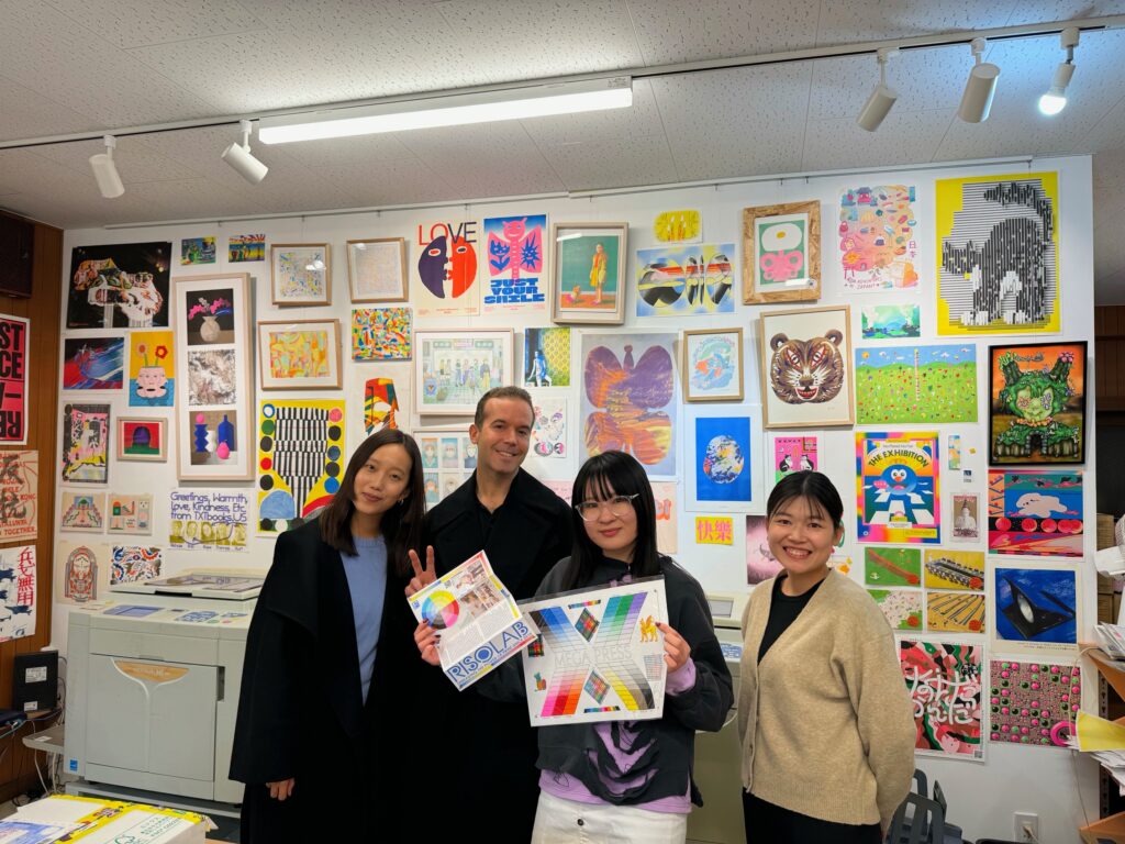 Three women and one man standing and smiling holding prints in front of a wall of prints