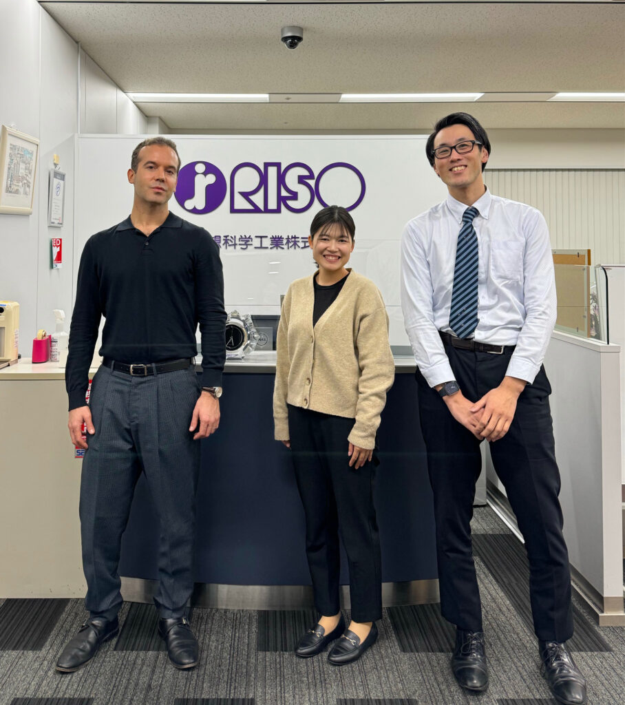 Two men and a woman standing in front of the Riso, Inc sign at Riso Kagaku HQ in Tokyo, Japan