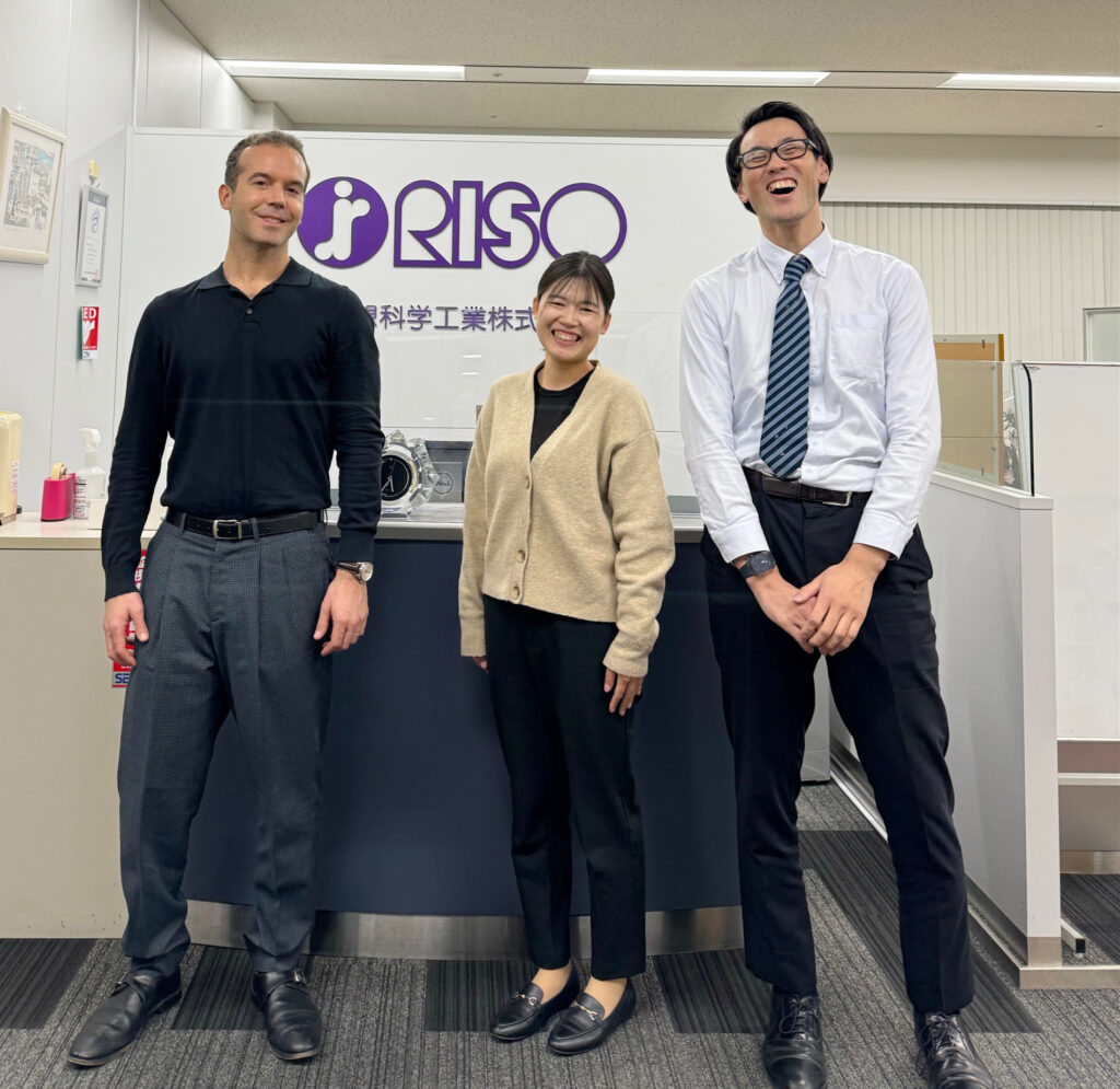 Two men and a woman standing in an office in front of the Riso sign