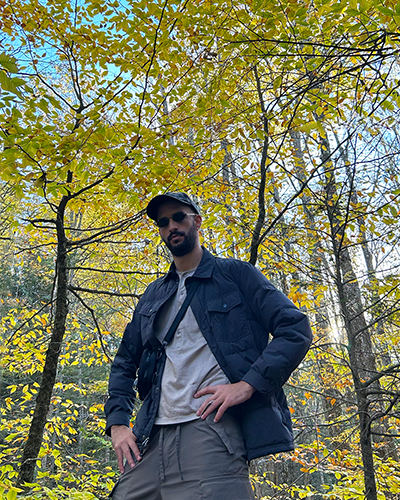 Photo of Chase Taylor, a white man with a beard and sunglasses, posing with a hand on the hip in a forest.