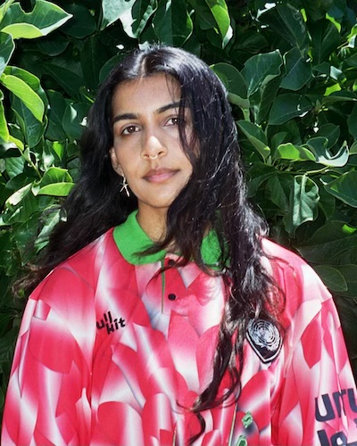 Headshot of Sarah Zarina Hakani, a South Asian woman with long, wavy dark hair, a septum piercing and wearing an oversized red and green jersey against a background of foliage.