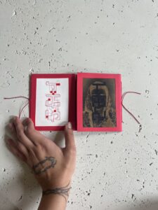 Open spread of Sarah Hakani Zarina's publication, an accordion book containing two single pamphlets with red paper frames, one side with black and red geometric designs printed on white paper, the other side with gold designs printed on black paper.