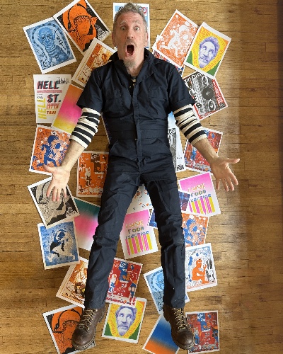 Full body photo of Josh Gosfield, a white man with gray hair and a goatee, wearing a black jumpsuit, work boots and long sleeved striped shirt underneath. He poses with his hands spread and feet apart with a shocked, open mouthed expression against a wooden background and his scattered colorful prints in the background.