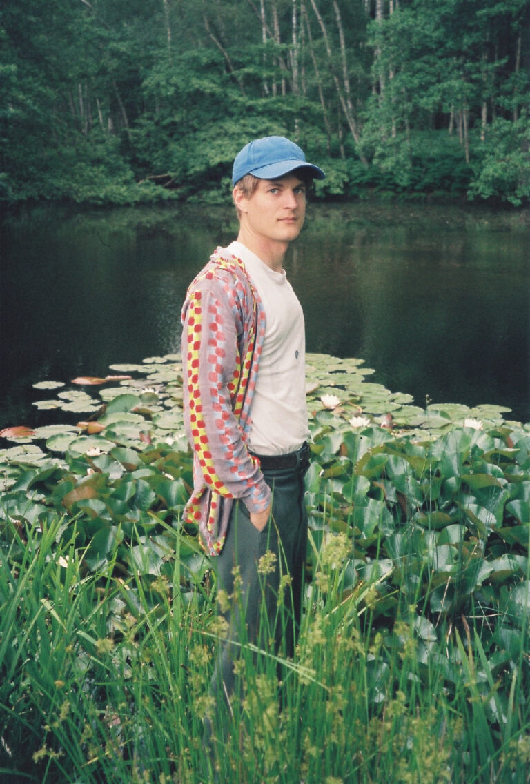 Photograph of Emil, Danish artist wearing a blue baseball hat, a patterned cardigan and standing in front of a pond with lily pads.