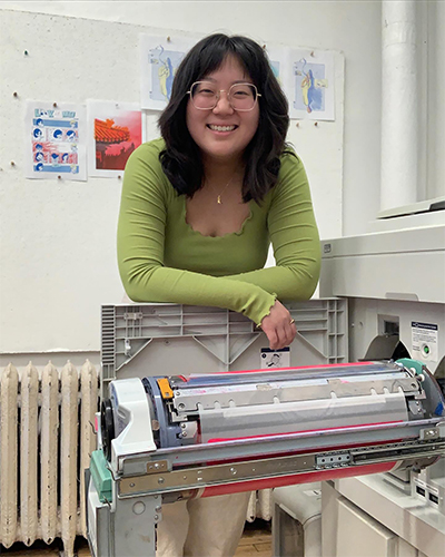 Photo of Suejee Lee, who poses with a riso printer. She wears glasses and has shoulder length wavy black hair.