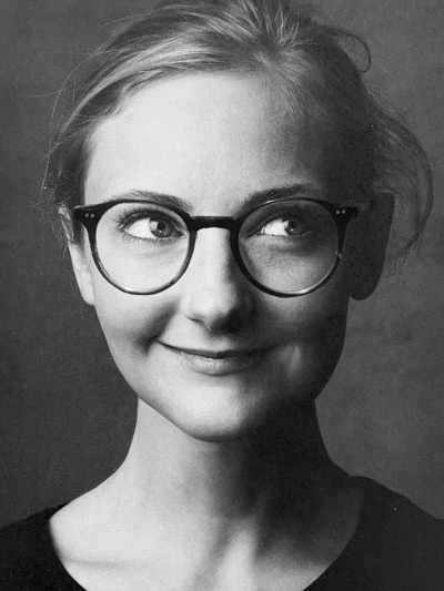 Black and white headshot of Kelli Anderson, she has light hair, round black glasses and looks off to her right.