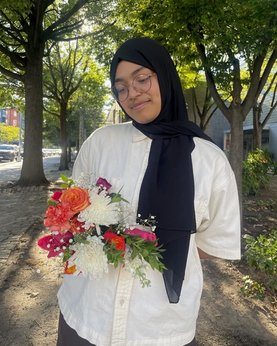Afrida Ahmed holds a bouquet of flowers, wears a white shirt, a hijab, glasses and has a septum ring.