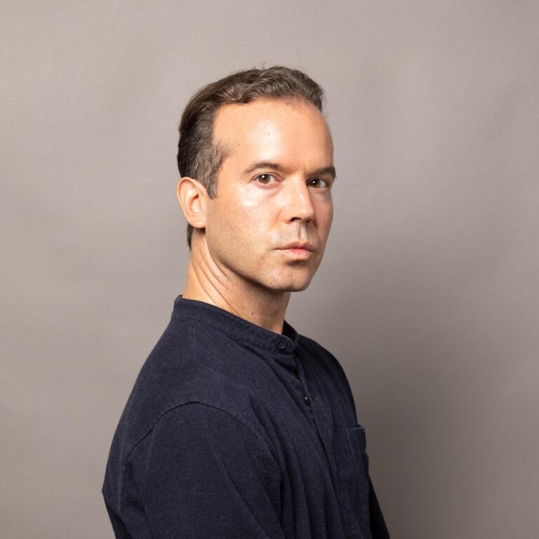 Middle aged man, slightly tan, looking towards the camera with three quarters profile, grey background, blue shirt, strong gaze