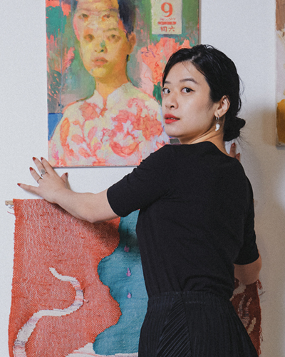 Singaporean Artist Jia Sung hangs up her paintings in her studio, looking over her shoulder. She has her hair in a bun, wears black and bright red lipstick.