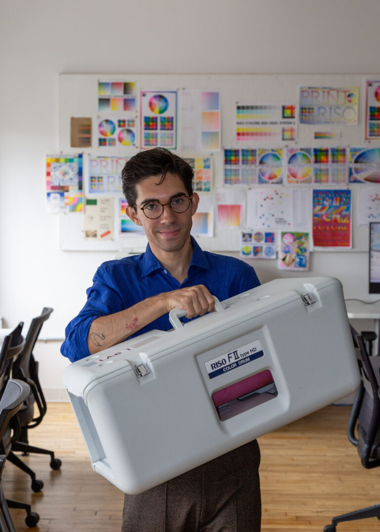 Photo of Kyle at the RisoLAB, he has glasses, dark hair, wears a blue shirt and is holding a riso drum case.